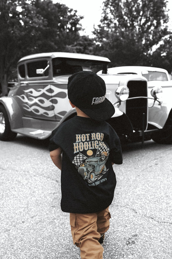 Child wearing a 'Hot Rod Hooligan' t-shirt and cap, standing in front of vintage cars.